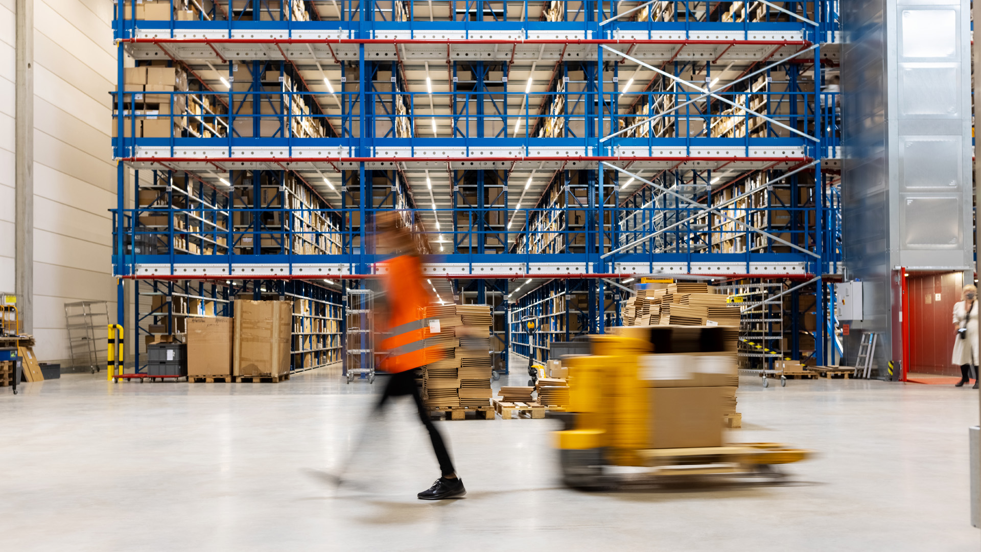 Blurry image of person wearing orange safety vest walking quickly through a factory with scaffolding and shelving in the background